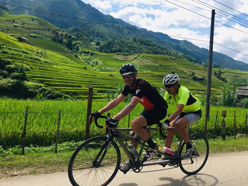Ashley piloting Lindy at the front of a tandem bike cycling in north-west Vietnam with views of rice terraces behind them