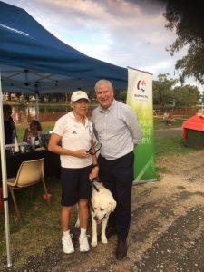 Lindy and Michael McCormack MP with Comet standing in front of a tent and Australia Day banner with the river behind