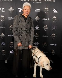 Lindy dressed up in jacket standing with Comet in harness beside her in front of the sponsor board for the Paralympics Australia Awards