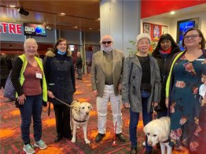 Group of six people, two with guide dogs and three audio description volunteers standing in the foyer of the Canberra Theatre