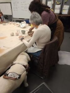 Comet standing beside Lindy who is seated with a ceramic object in front of her being assisted by a lady at a hands-on gallery workshop